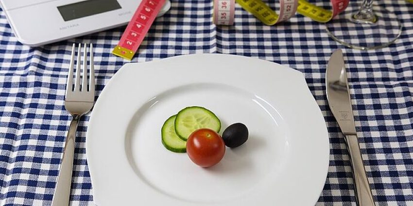 Ein Teller mit zwei Scheiben Gurken einer Tomate und einer Olive, daneben Messer und Gabel darüber eine Waage und ein Massband