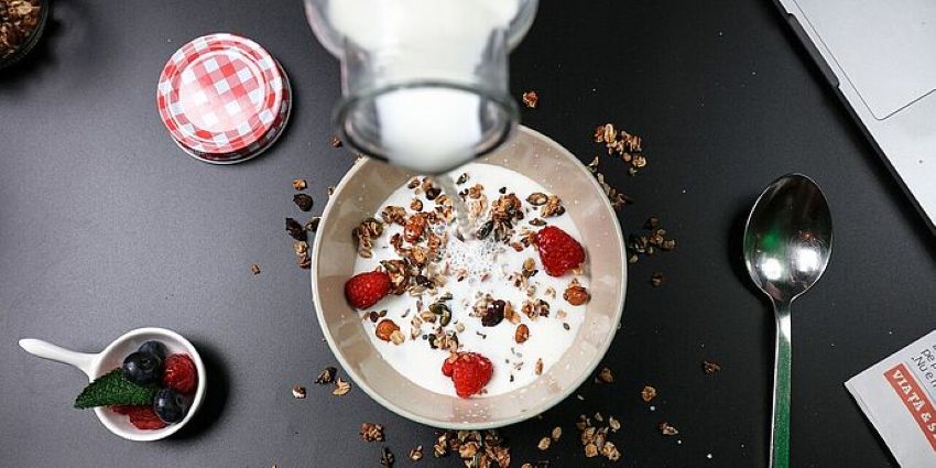 Müsli auf einem Tisch in das soeben Milch gegossen wird, daneben eine kleine Schale mit Früchten und ein Löffel