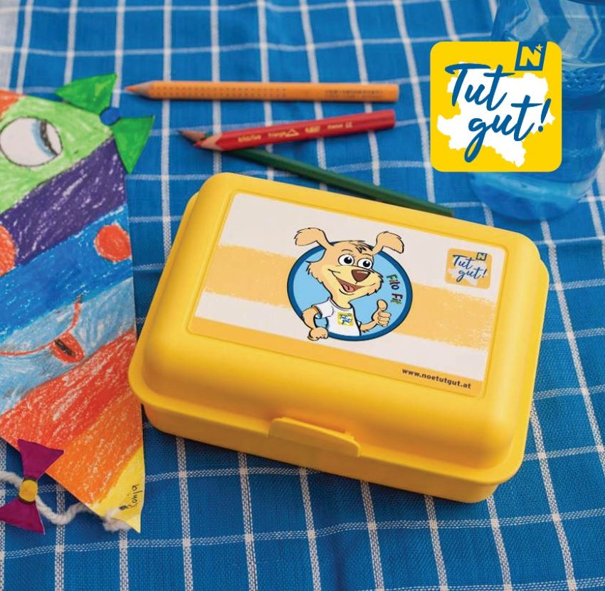 Eine Jausenbox mit Fito Fit, ein gebastelter Drachen und Buntstifte auf einem blau karierten Tischtuch