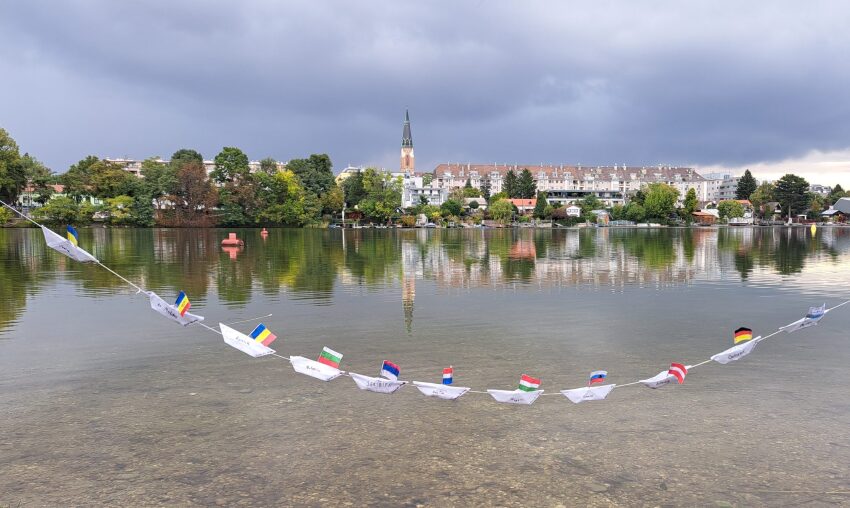 Fluss mit einer Stadt im Hintergrund, vielen Bäumen und darüber ist eine Schnur mit Wimpel darauf gespannt - auf diesen Wimpel sieht man Flaggen verschiedener Nationen