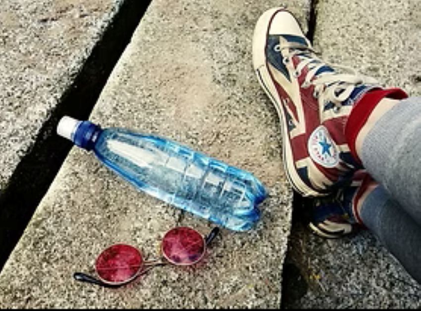am Boden liegt eine Trinkflasche mit Wasser gefüllt sowie eine Sonnenbrille und daneben eine Person, von der nur die Schuhe zu sehen sind