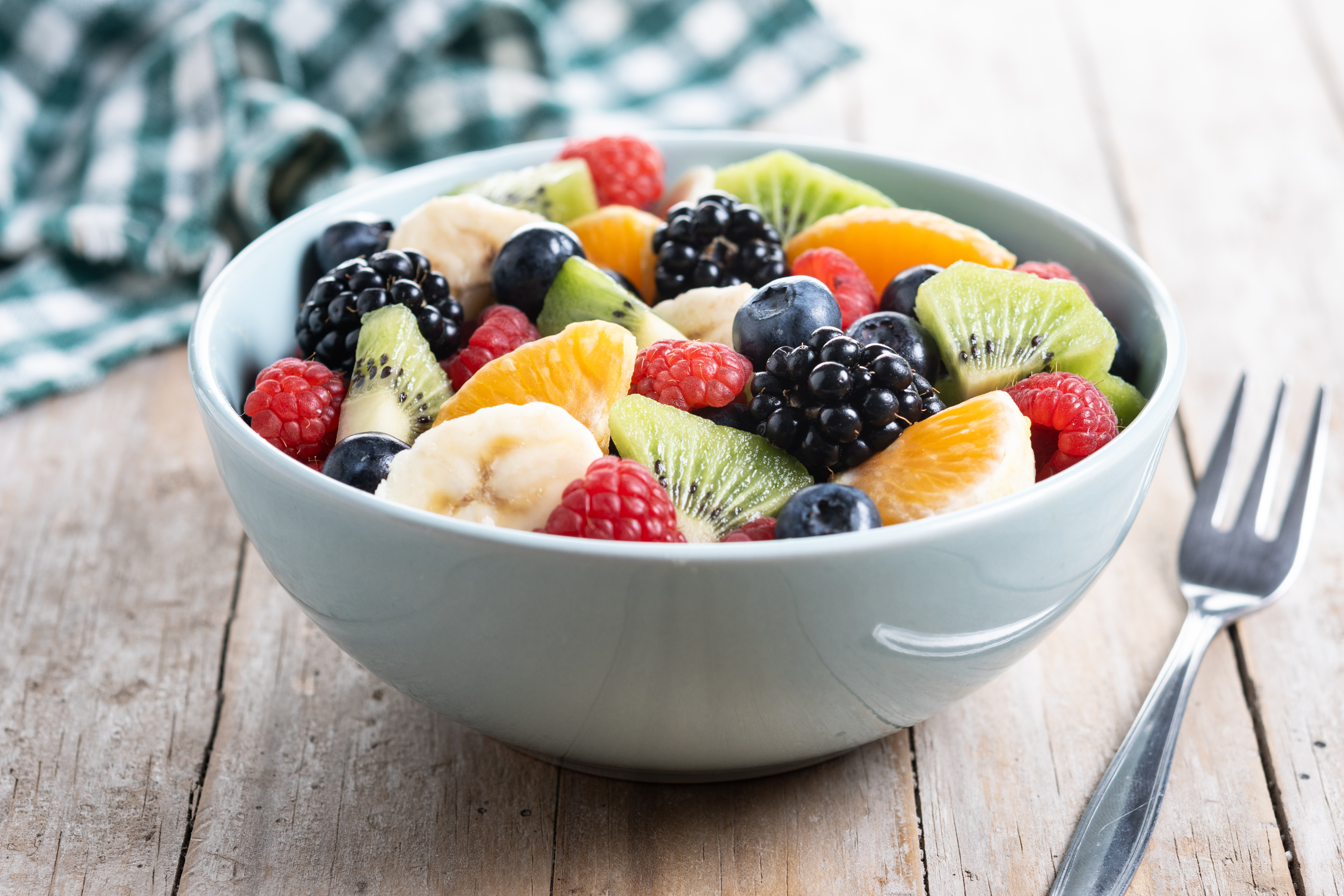 bunter Fruchtsalat mit Kiwi, Banane, Himbeere, Schwarzbeere, Orange in einer blauen Schüssel auf einem Holztisch